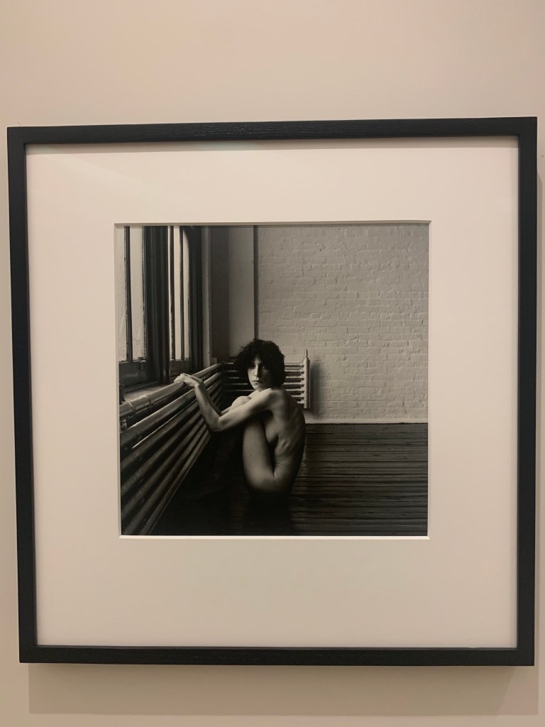 black and white photo of patti smith nude and crouched down on her toes holding a furnace in a bare white brick apartment
